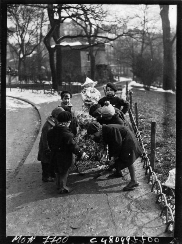 Children are gathered around and building a snowman on a snowy pathway, with trees and a large building in the background. Some hold snow and sticks to add to the snowman, and the ground is partially covered with snow.