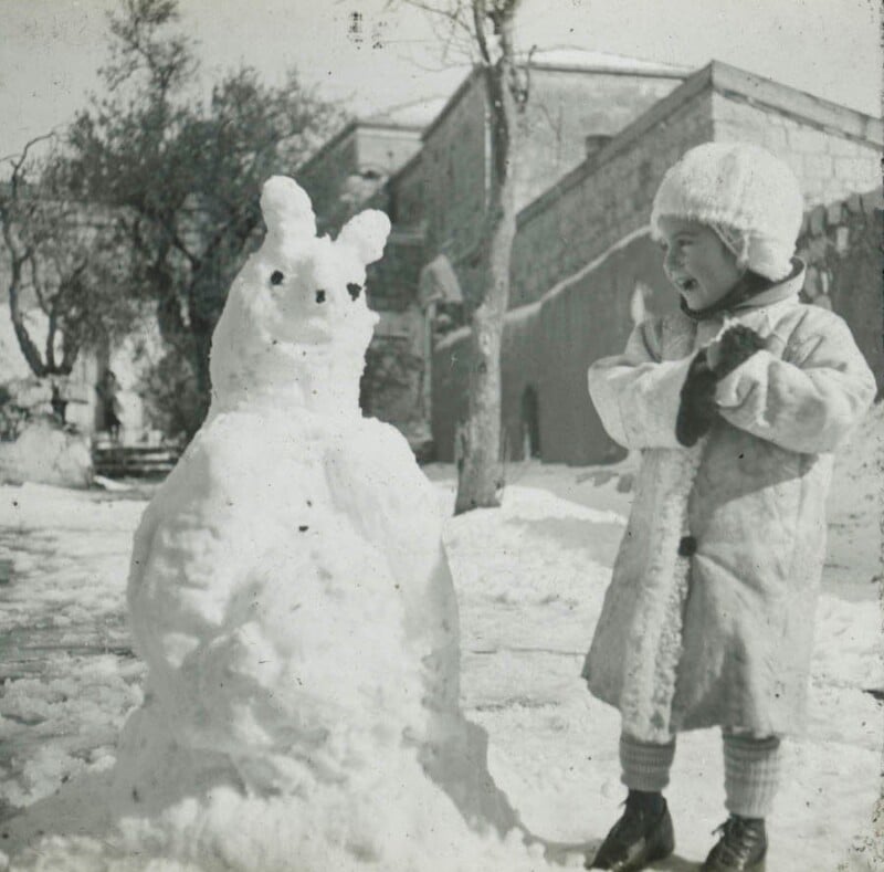 A child in a winter coat and hat stands beside a snowman with twig arms and two lumps for ears. The snowman is in a snowy courtyard with trees and a building in the background. The child appears to be smiling.