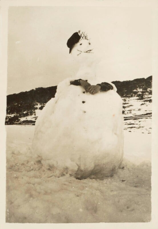 A vintage photo of a snowman wearing a black hat. The snowman has a small mouth and stick arms with gloves. It stands on a snowy landscape with hills in the background.