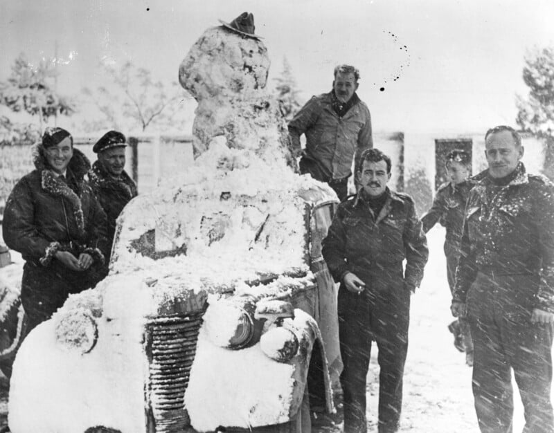 Five men wearing winter clothing stand beside a snow-covered car, which has a snowman wearing a hat on its hood. Snow blankets the ground and trees in the background, creating a wintry scene.