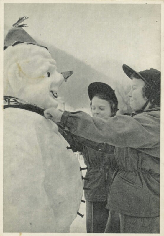 Two children in winter coats and hats build a snowman, adding buttons and adjusting its scarf. The snowman wears a pointed hat and has a carrot nose. Snow covers the ground, with mountains visible in the background.