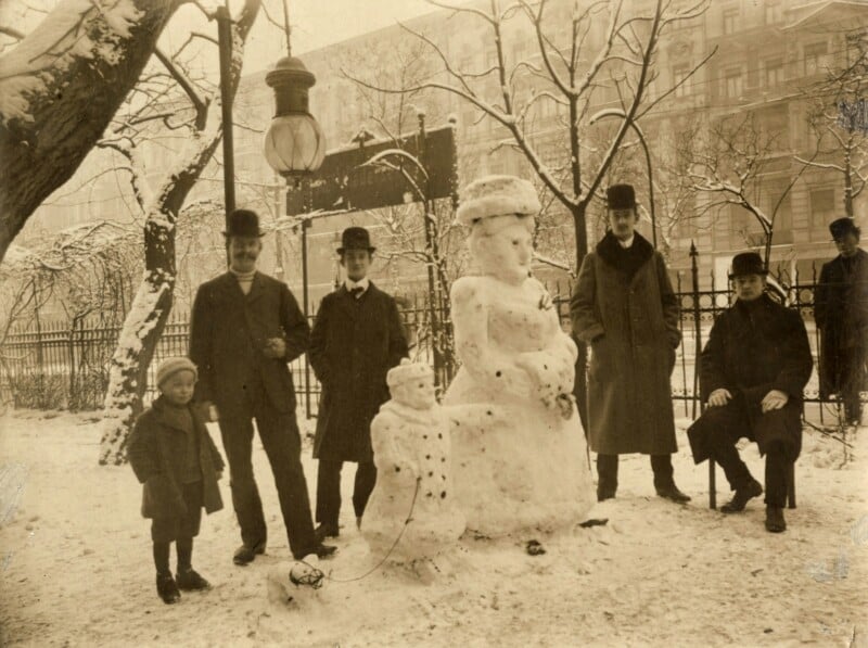 Vintage photo of six people standing around a snowwoman and snowdog in a snowy park. They are dressed in early 20th-century clothing, with men in coats and bowler hats, and a child in a coat and hat. Snow-covered trees and a lamppost are in the background.