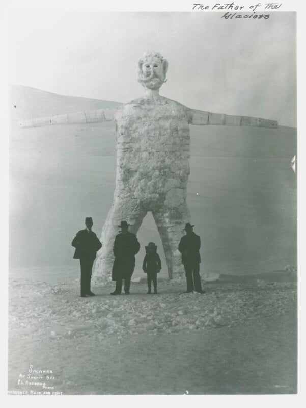 Four people stand in front of a large snowman sculpture resembling a human figure with outstretched arms. The snowman has a carved face and legs spread apart. The environment is snowy, and a handwritten note reads "The Father of the Glaciers.