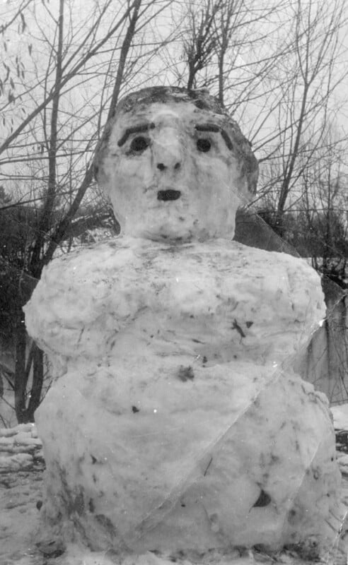 A large snowman with a detailed face, including eyes and a mouth, stands in a snowy area. Trees with bare branches are visible in the background. The snowman has rounded, bulky arms and a textured surface.