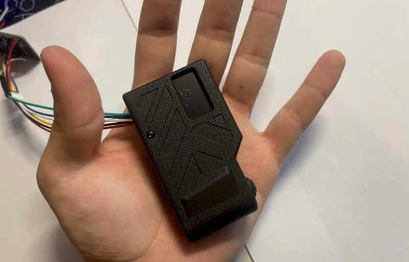 A person holds a rectangular black electronic device in their hand. The device has a textured surface with a geometric pattern and multiple wires extending from one side.