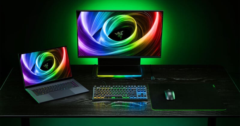 A gaming setup on a black desk featuring a laptop, a large monitor displaying vibrant multicolored swirls, a mechanical keyboard with RGB lighting, a black mouse, and a large mouse pad. Green ambient lighting illuminates the scene.