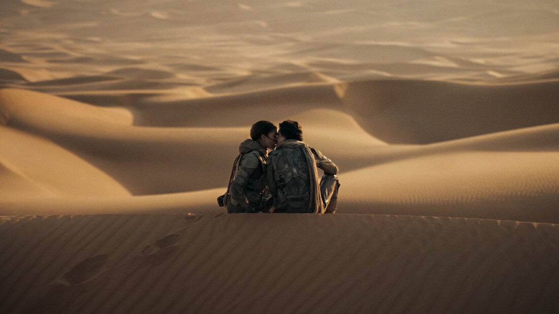 Two people, wearing backpacks, sit closely together on a sunlit sand dune in a vast desert landscape. The sand is smooth, with gentle ridges and footprints leading up to where they rest, creating a serene scene.