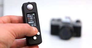 Close-up of a hand holding a black light meter with a display showing EV, shutter speed, aperture, and color temperature readings. A blurred camera is visible in the background.