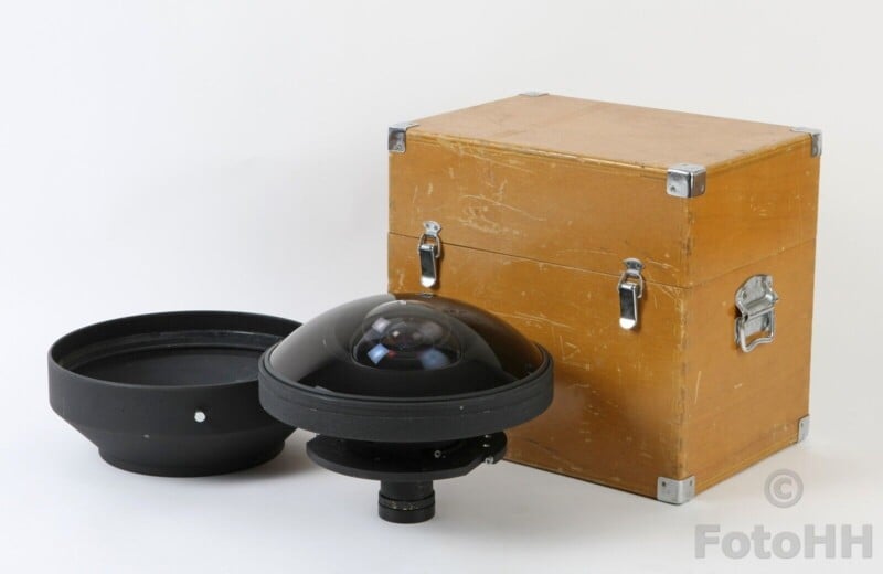 A fisheye camera lens with a dome-shaped glass cover and a black base is positioned next to a large, closed wooden storage box with metal corner protectors and latches. The lens cap is placed beside the lens.