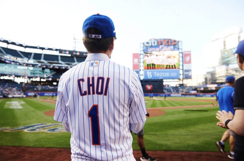 An individual wearing a baseball jersey displaying the name "CHOI" and the number "1" standing by the edge of a baseball field. The stadium basking in sunlight, and the scoreboard in the backdrop highlights a group of players.