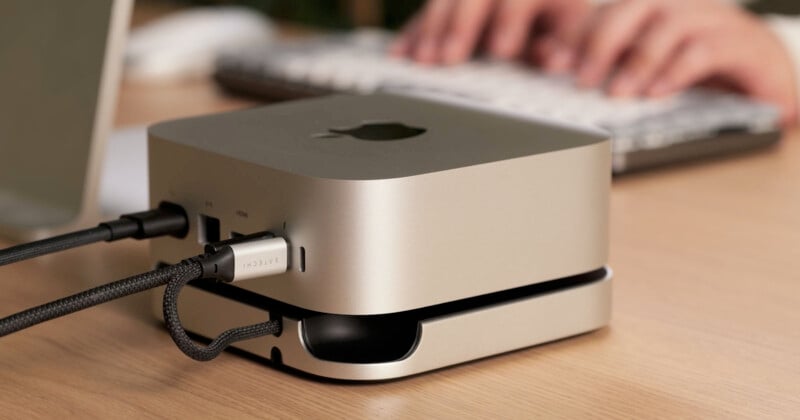 A compact computer with a metallic finish is on a desk, connected to cables. In the background, blurred hands type on a keyboard.