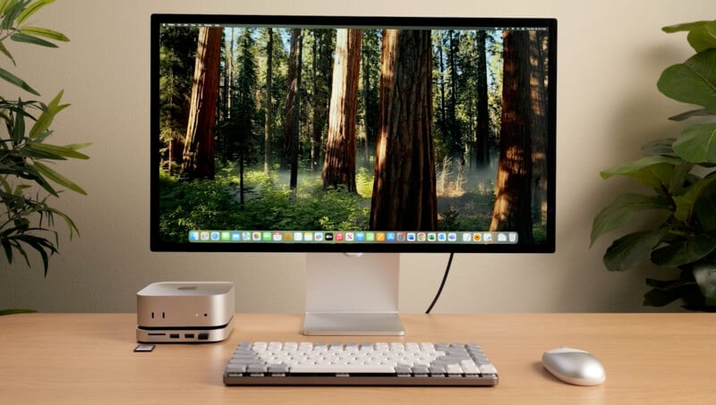 A desktop setup featuring a monitor displaying a forest scene, a compact silver computer, a white and gray mechanical keyboard, and a wireless mouse. The background has a light wall with green plants on the sides.