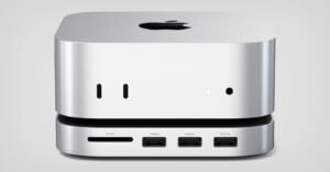 A compact silver computer with an Apple logo on top is shown from the front. It features ports for SD cards, USB-C, HDMI, and USB 2.0 connections. The device has a sleek, minimalist design.