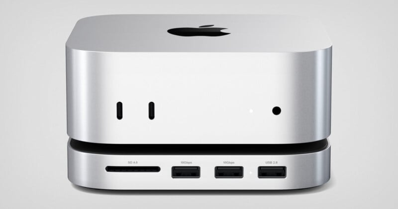 A compact silver computer with an Apple logo on top is shown from the front. It features ports for SD cards, USB-C, HDMI, and USB 2.0 connections. The device has a sleek, minimalist design.