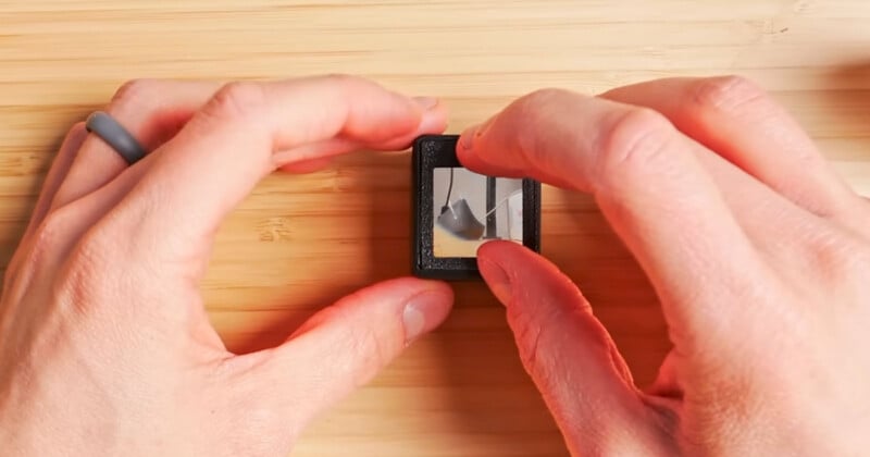 Two hands manipulate a small, reflective prism on a wooden surface. The prism shows the reflection of a nearby black object, possibly a camera lens. A person is wearing a gray ring on their right hand.