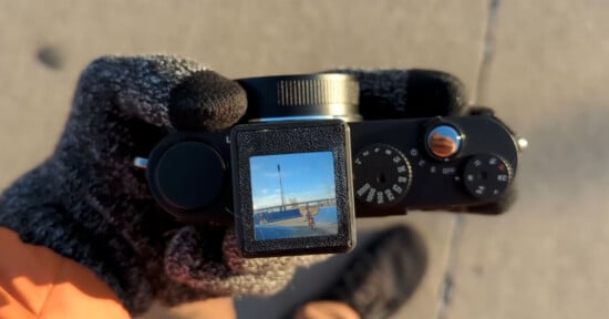 Gloved hand holding a camera outdoors, with a display screen showing a vibrant image of a skateboarder performing a trick on a ramp under a clear blue sky. The camera's dials and controls are visible.