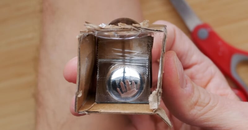 A person is holding a small cardboard box. Inside, there is a shiny metallic sphere. In the background, there is a pair of red-handled scissors on a wooden surface.