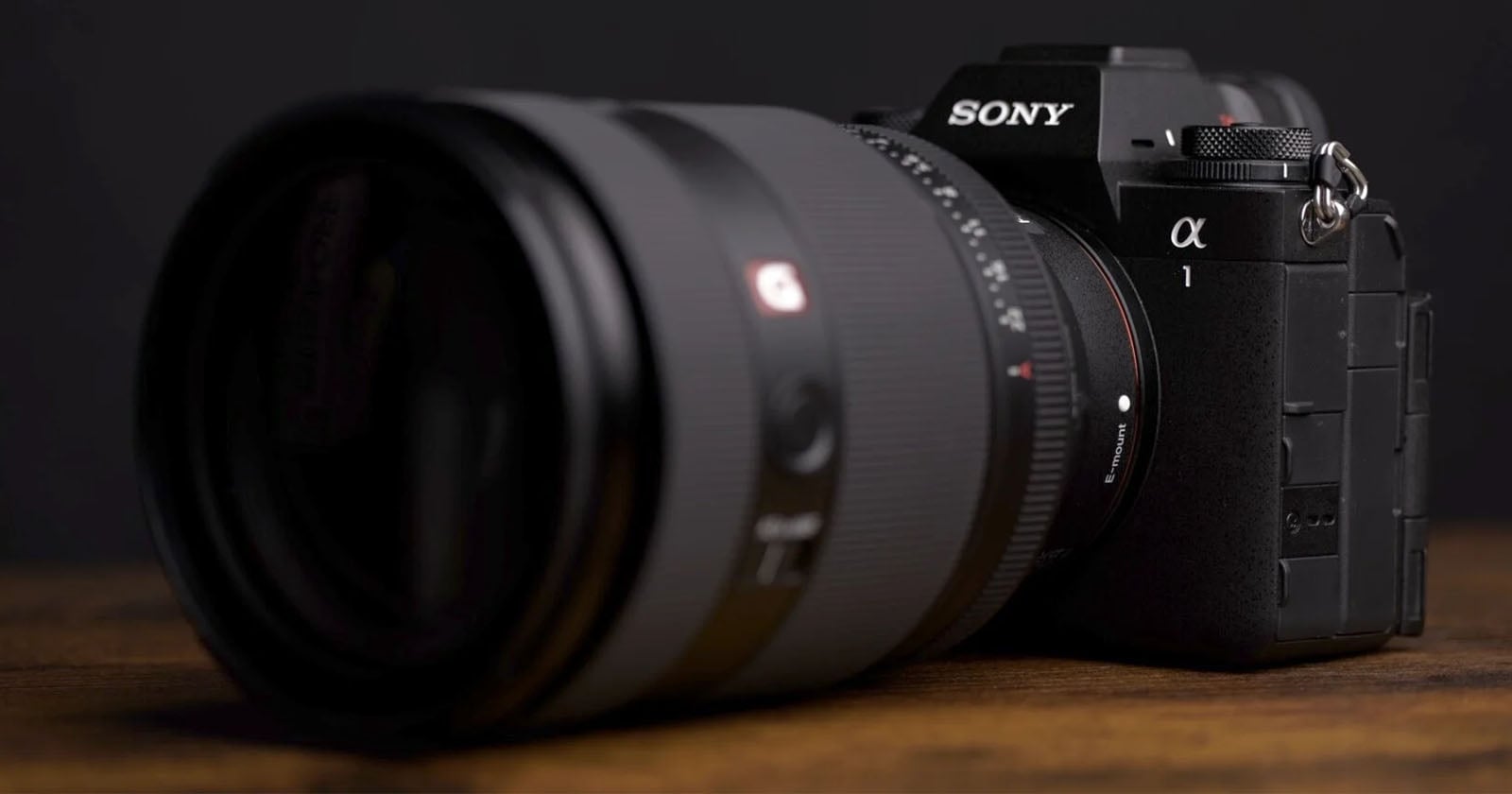 A Sony Alpha camera with a large lens is placed on a wooden surface. The focus is on the lens, while the camera body features prominent branding. The background is dark, highlighting the camera's sleek design.