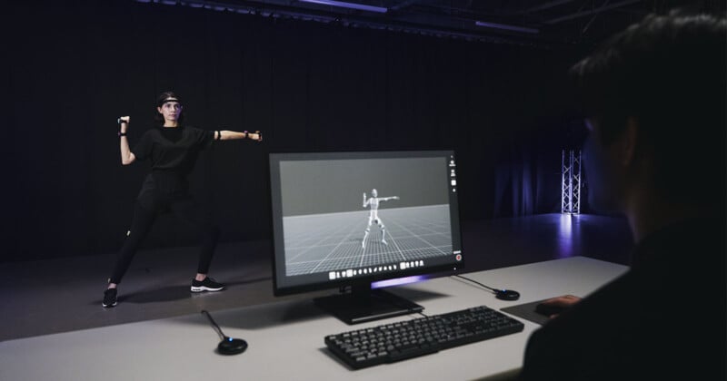 A person wearing black motion capture equipment performs in front of a camera in a dark room. A computer monitor on a desk displays a digital skeleton mimicking their pose.