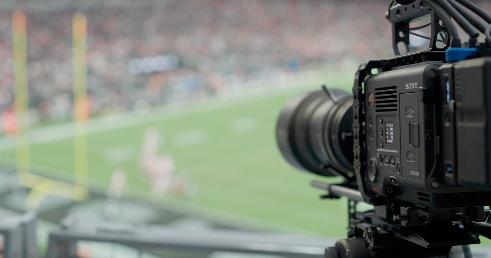 A close-up of a professional video camera filming an American football game. The camera is positioned to capture action on the field with blurred players and goalposts visible in the background, indicating focus on sports broadcasting.