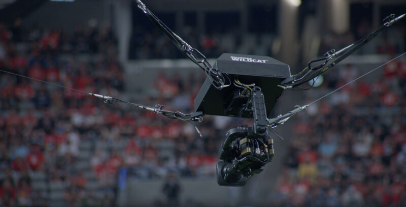 A suspended camera system labeled "WILDCAT" is set up on wires above a sports stadium, capturing the game. The blurred audience in red attire fills the background, indicating a lively sporting event.