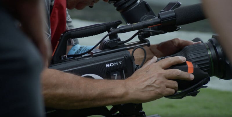 A close-up of a person operating a large Sony video camera. The person is adjusting the lens while looking through the viewfinder, capturing footage outdoors. The scene suggests a professional filming setting.