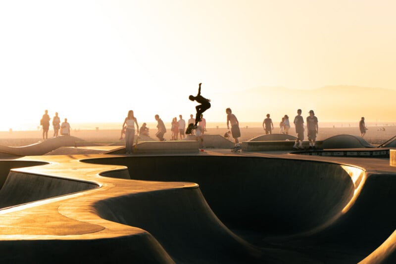 A skateboarder executes a trick in mid-air at a skatepark during sunset. The sky appears hazy, casting a warm hue over the environment. Silhouetted figures are positioned around the park, with distant hills observable in the background.