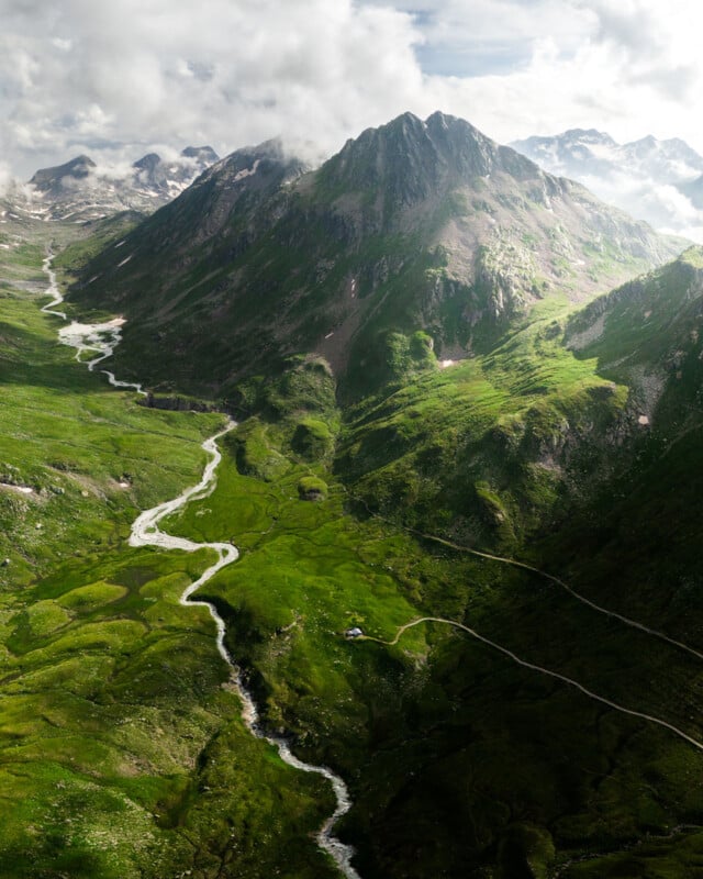 Bird's-eye view of a vibrant, green mountainous landscape with a meandering river traversing the valley. The rugged peaks are partly cloaked in clouds, and the scene is bathed in gentle, natural illumination.