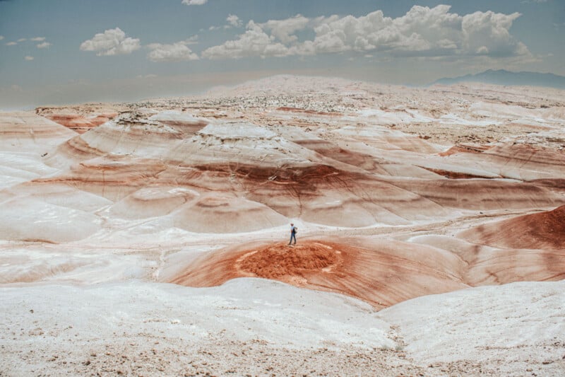 An individual stands solitary on an expansive, colorful desert landscape showcasing undulating hills in shades of red, brown, and white beneath a cloudy sky. The view is vast and isolated, with distant peaks visible in the background.