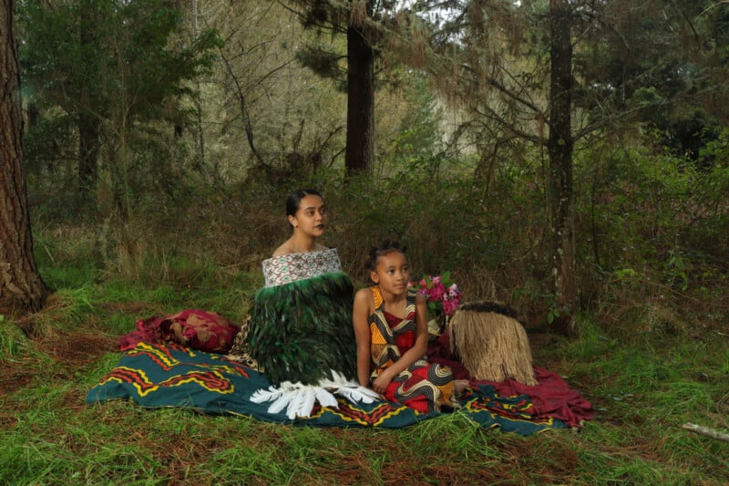 Two individuals, clad in traditional garments adorned with feathers and woven designs, sit on an embellished cloth within a verdant forest setting. Surrounding them is lush greenery, and a handcrafted basket with flowers rests nearby.