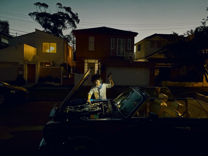 An individual inspects the engine of a vehicle at night while utilizing a flashlight. The vehicle is parked on a residential avenue with modern homes in the backdrop, some illuminated. The atmosphere is dim, indicating later evening or nighttime.