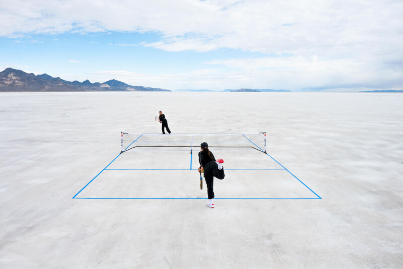 Two people play tennis on a minimalistic court​ marked⁢ with blue lines⁢ on a ⁤vast, flat ‍salt plain. The sky is partly cloudy with distant mountains in the background, creating a surreal landscape.