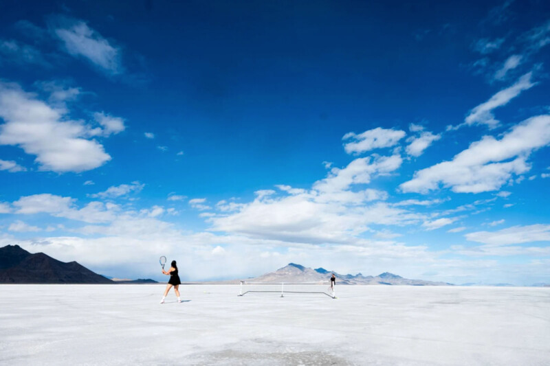 Two tennis players⁢ compete on a vast, flat white‌ surface under a bright blue sky with scattered ⁤clouds. Mountains rise in the distance, adding a surreal touch to the scene.