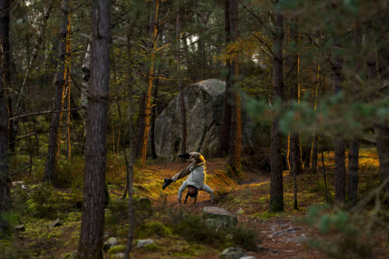 A breakdancer performs a handstand in a dense forest, surrounded by trees and rocks, with sunlight filtering through the branches.