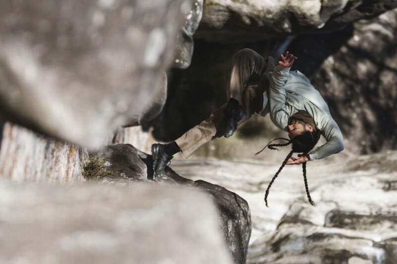 A person‌ with braids, wearing a light-colored ⁤jacket and beige pants, gracefully performs an acrobatic move ‍on large, gray rocks,⁤ displaying agility and strength. The perspective ​highlights the rocky environment.