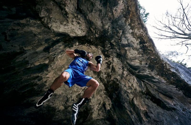 A boxer in blue shorts and black gloves is captured mid-air against a rocky cave ceiling. The perspective is from below, emphasizing the height and texture of the rocks. Bare tree branches are visible at the opening above.