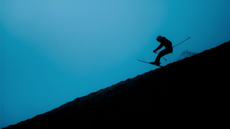 Silhouette of a person skiing downhill against a blue sky, angled sharply to the right. The skier is in mid-action, with poles outstretched and snow spraying behind, creating a dynamic sense of movement.