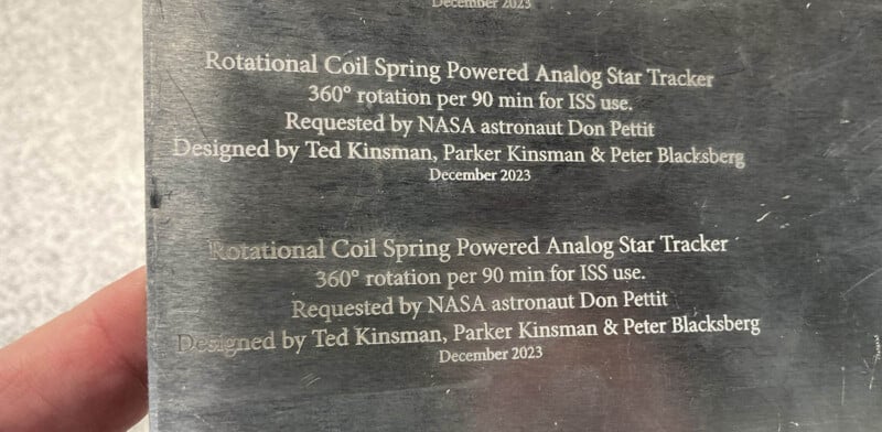 Plaque displaying text about a Rotational Coil Spring Powered Analog Star Tracker, with specifications for ISS use. It mentions NASA astronaut Don Pettit and designers Ted Kinsman, Parker Kinsman, and Peter Blacksberg. Dated December 2023.