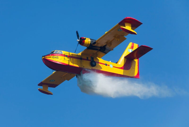 A yellow and red amphibious aircraft releases water while flying against a clear blue sky, used for firefighting operations.