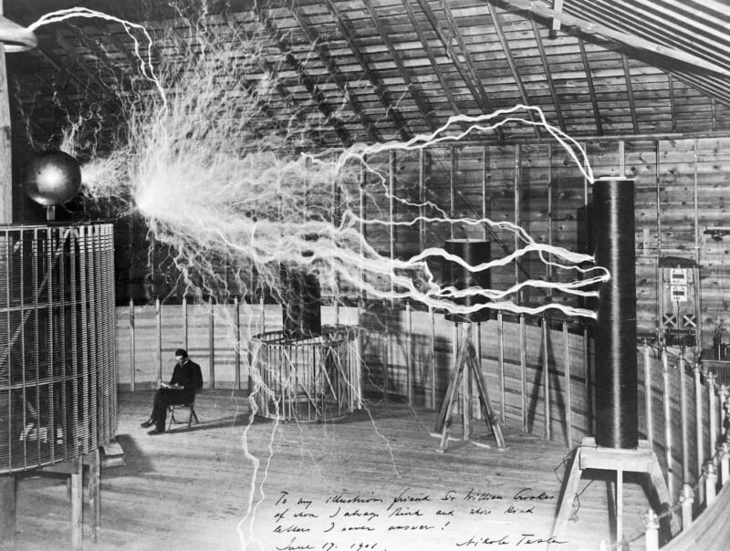 A black-and-white photo of a tesla coil indoors, emitting large electrical arcs. A person is seated nearby reading or writing. The wooden walls and metal equipment are visible in the spacious room. Handwritten text is on the bottom of the image.