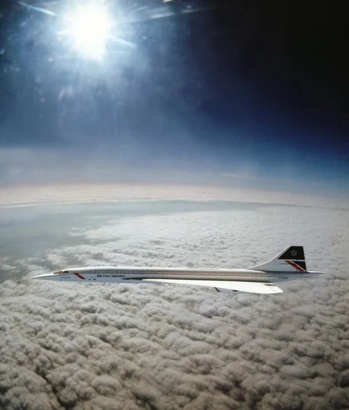 An airplane with a sleek, pointed nose flies above a layer of fluffy clouds. The sun shines brightly in the sky, casting a glow over the aircraft. "British Airways" is visible on the side of the plane.