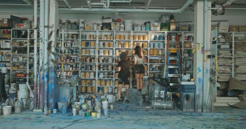 Two people stand on stools, facing a wall of shelves filled with various paint cans and supplies in a messy, colorful workshop. The floor is covered with splatters, and various tools and containers are scattered around.