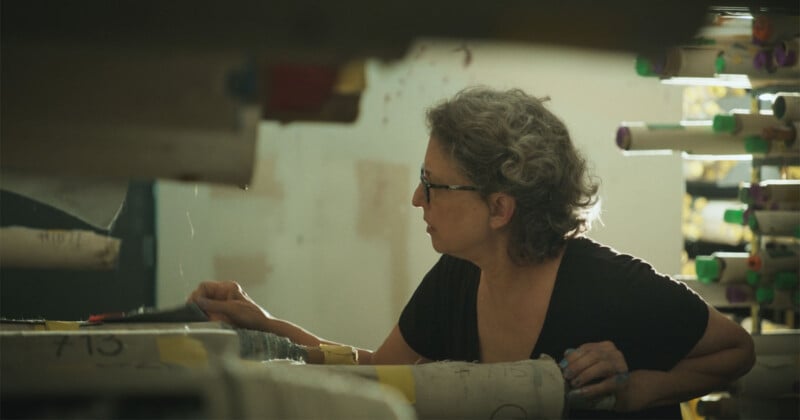 A person with curly gray hair and glasses is examining fabric rolls. They are wearing a black shirt and focused on their task in a workshop setting, with various colored fabric cylinders around. Light is casting warm tones across the scene.