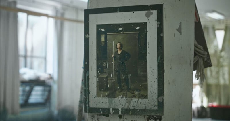 A framed photograph on a concrete wall shows a person standing confidently in a room. The surrounding area is dimly lit, with some clutter visible. Light filters in from windows in the background, adding a soft glow.