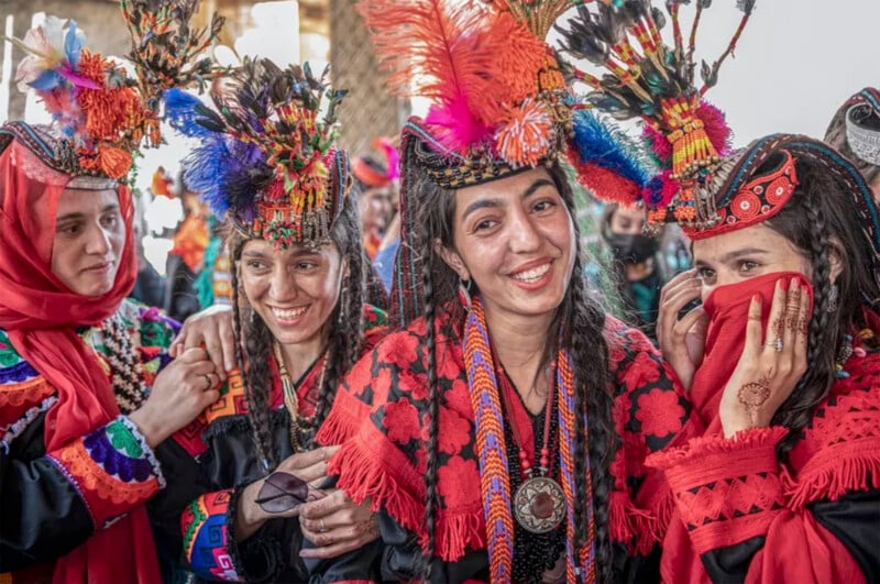 A group of women in vibrant traditional attire and ornate headpieces smile and interact. They wear colorful dresses with intricate patterns, one holding a pair of sunglasses, and another covering her face with a red scarf, displaying henna designs.