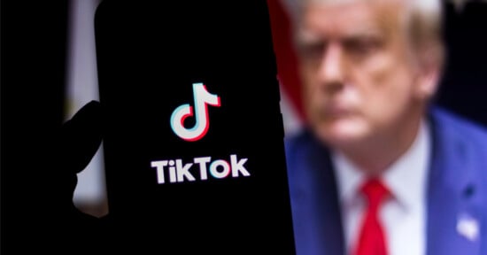 A smartphone displays the TikTok logo on its screen. In the blurred background, a person in a suit with a red tie is visible.