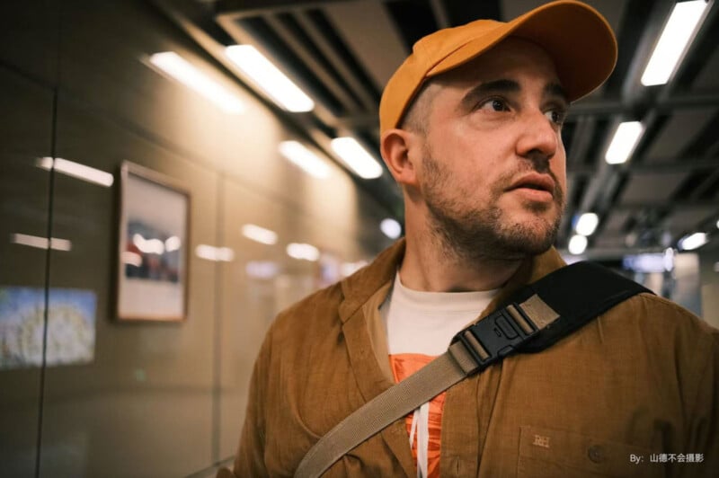 A man with a beard and a yellow cap looks to the side while standing in a modern indoor setting with bright overhead lights. He is wearing a brown jacket and carrying a shoulder bag.