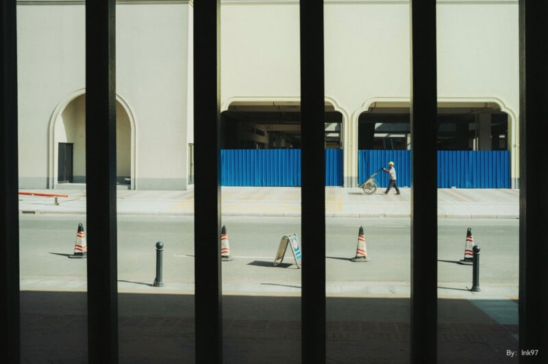 A person wearing a blue shirt and hat pushes a cart across an empty street, framed by dark vertical bars. Blue fencing and beige building in the background. Orange traffic cones line the street.