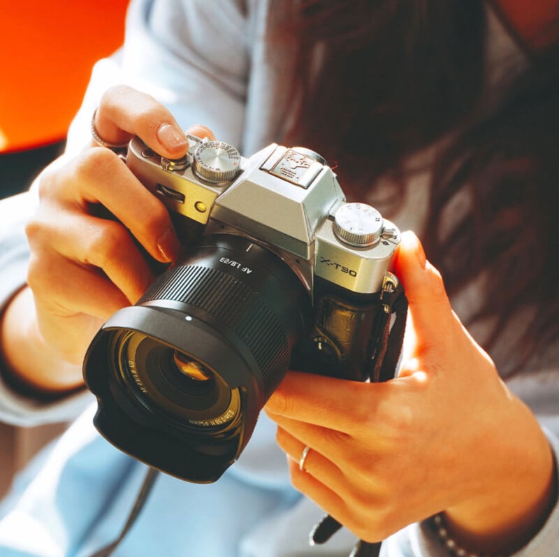 A person holding a silver and black camera with both hands, adjusting the lens. The camera has visible dials and a strap, and the background is softly blurred.