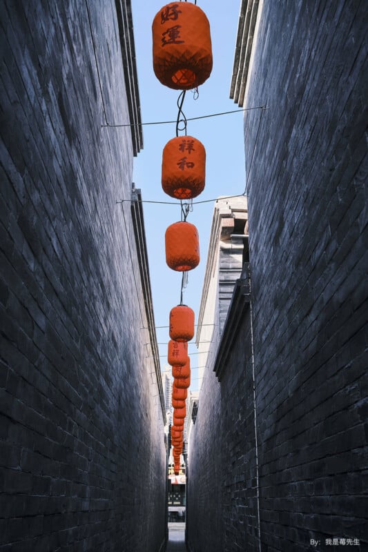 A narrow alleyway with tall brick walls features a string of vibrant red lanterns hanging overhead. The lanterns have Chinese characters on them and contrast against the blue sky visible between the walls.